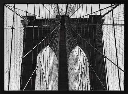 A low-angle, black and white photograph looking up at the iconic stone towers of the Brooklyn Bridge. The intricate web of suspension cables and vertical suspender ropes creates a symmetrical, geometric pattern against the light sky. The photo is enclosed in a silver frame. Print
