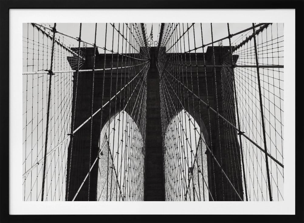 A low-angle, black and white photograph looking up at the iconic stone towers of the Brooklyn Bridge. The intricate web of suspension cables and vertical suspender ropes creates a symmetrical, geometric pattern against the light sky. The photo is enclosed in a silver frame. Print