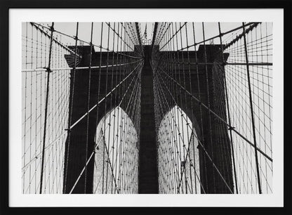 A low-angle, black and white photograph looking up at the iconic stone towers of the Brooklyn Bridge. The intricate web of suspension cables and vertical suspender ropes creates a symmetrical, geometric pattern against the light sky. The photo is enclosed in a silver frame. Print