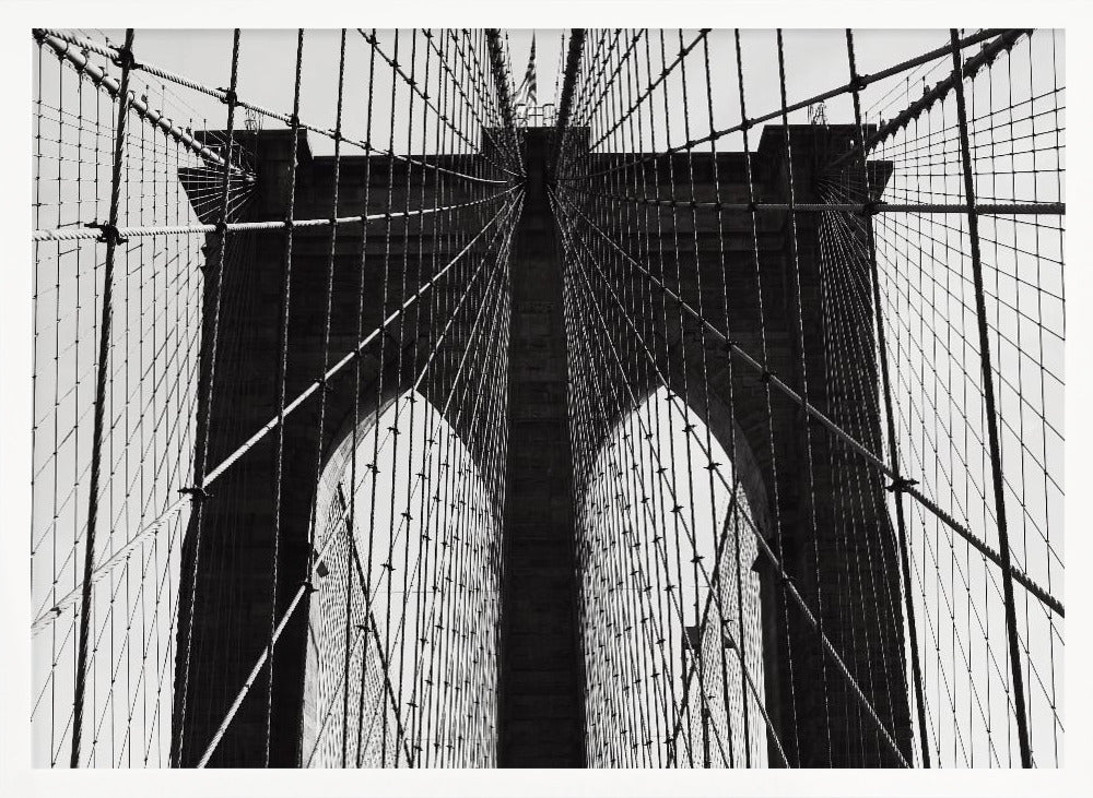 A low-angle, black and white photograph looking up at the iconic stone towers of the Brooklyn Bridge. The intricate web of suspension cables and vertical suspender ropes creates a symmetrical, geometric pattern against the light sky. The photo is enclosed in a silver frame. Print