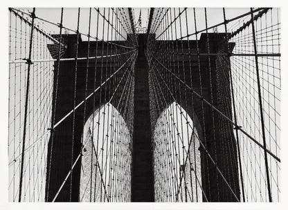 A low-angle, black and white photograph looking up at the iconic stone towers of the Brooklyn Bridge. The intricate web of suspension cables and vertical suspender ropes creates a symmetrical, geometric pattern against the light sky. The photo is enclosed in a silver frame. Print