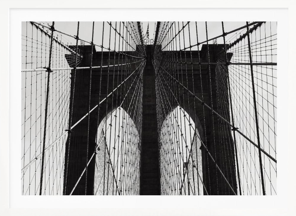 A low-angle, black and white photograph looking up at the iconic stone towers of the Brooklyn Bridge. The intricate web of suspension cables and vertical suspender ropes creates a symmetrical, geometric pattern against the light sky. The photo is enclosed in a silver frame. Print