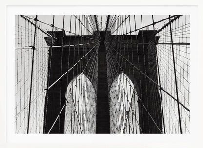 A low-angle, black and white photograph looking up at the iconic stone towers of the Brooklyn Bridge. The intricate web of suspension cables and vertical suspender ropes creates a symmetrical, geometric pattern against the light sky. The photo is enclosed in a silver frame. Print