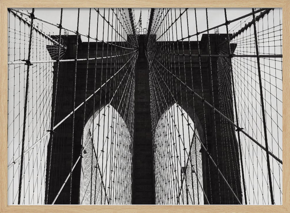 A low-angle, black and white photograph looking up at the iconic stone towers of the Brooklyn Bridge. The intricate web of suspension cables and vertical suspender ropes creates a symmetrical, geometric pattern against the light sky. The photo is enclosed in a silver frame. Print