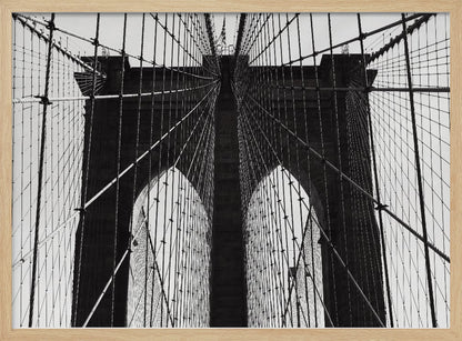 A low-angle, black and white photograph looking up at the iconic stone towers of the Brooklyn Bridge. The intricate web of suspension cables and vertical suspender ropes creates a symmetrical, geometric pattern against the light sky. The photo is enclosed in a silver frame. Print