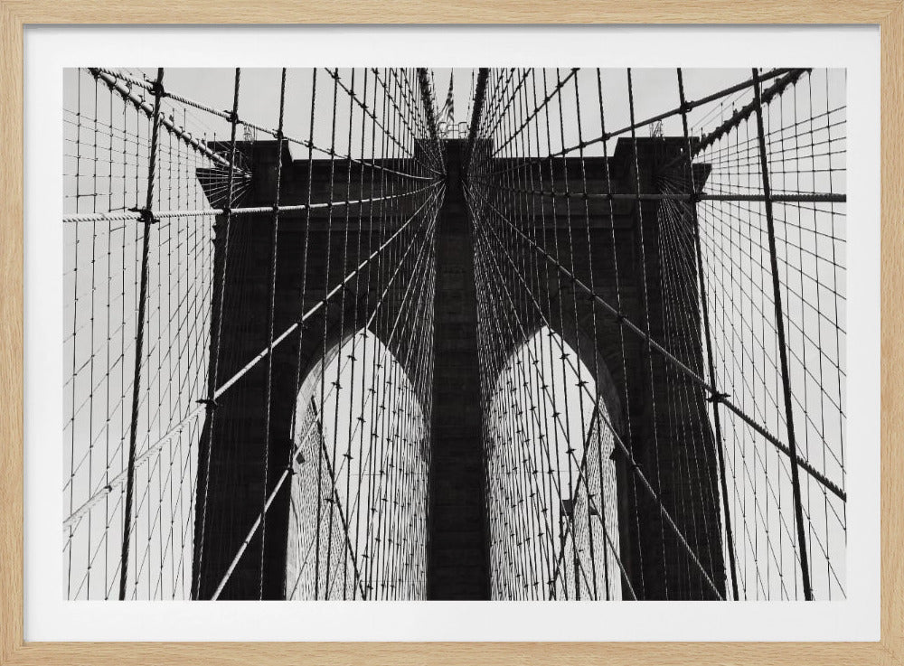 A low-angle, black and white photograph looking up at the iconic stone towers of the Brooklyn Bridge. The intricate web of suspension cables and vertical suspender ropes creates a symmetrical, geometric pattern against the light sky. The photo is enclosed in a silver frame. Print