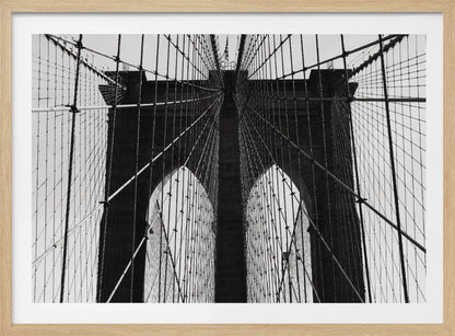 A low-angle, black and white photograph looking up at the iconic stone towers of the Brooklyn Bridge. The intricate web of suspension cables and vertical suspender ropes creates a symmetrical, geometric pattern against the light sky. The photo is enclosed in a silver frame. Print