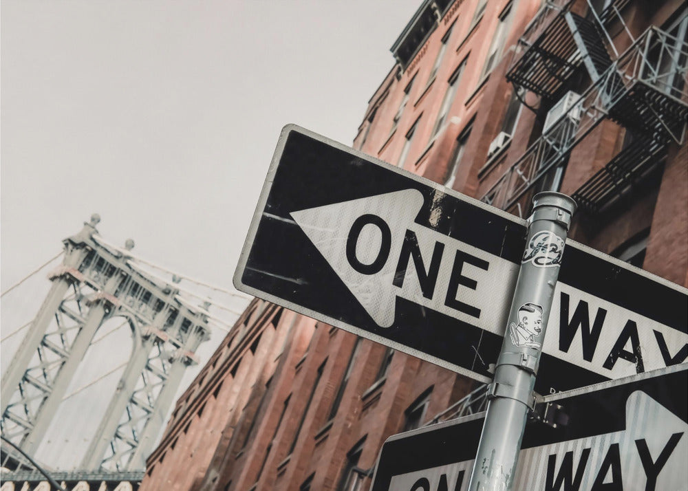 A low-angle shot of a black and white 'ONE WAY' street sign in the foreground, with a red brick building and the Manhattan Bridge visible in the background under an overcast sky, all enclosed in a silver frame. Artwork