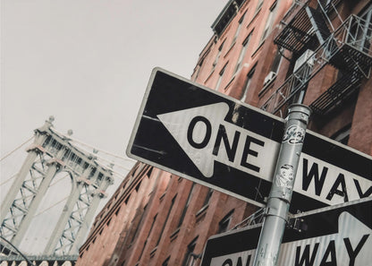 A low-angle shot of a black and white 'ONE WAY' street sign in the foreground, with a red brick building and the Manhattan Bridge visible in the background under an overcast sky, all enclosed in a silver frame. Artwork