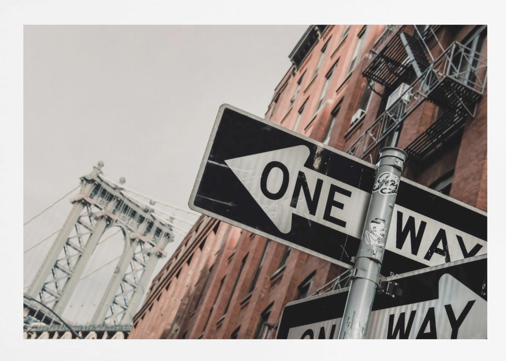 A low-angle shot of a black and white 'ONE WAY' street sign in the foreground, with a red brick building and the Manhattan Bridge visible in the background under an overcast sky, all enclosed in a silver frame. Artwork