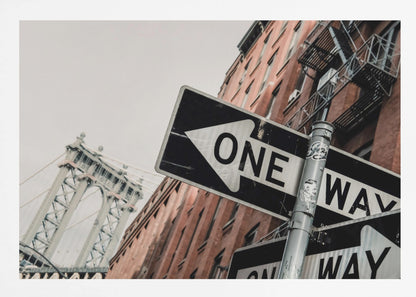 A low-angle shot of a black and white 'ONE WAY' street sign in the foreground, with a red brick building and the Manhattan Bridge visible in the background under an overcast sky, all enclosed in a silver frame. Artwork