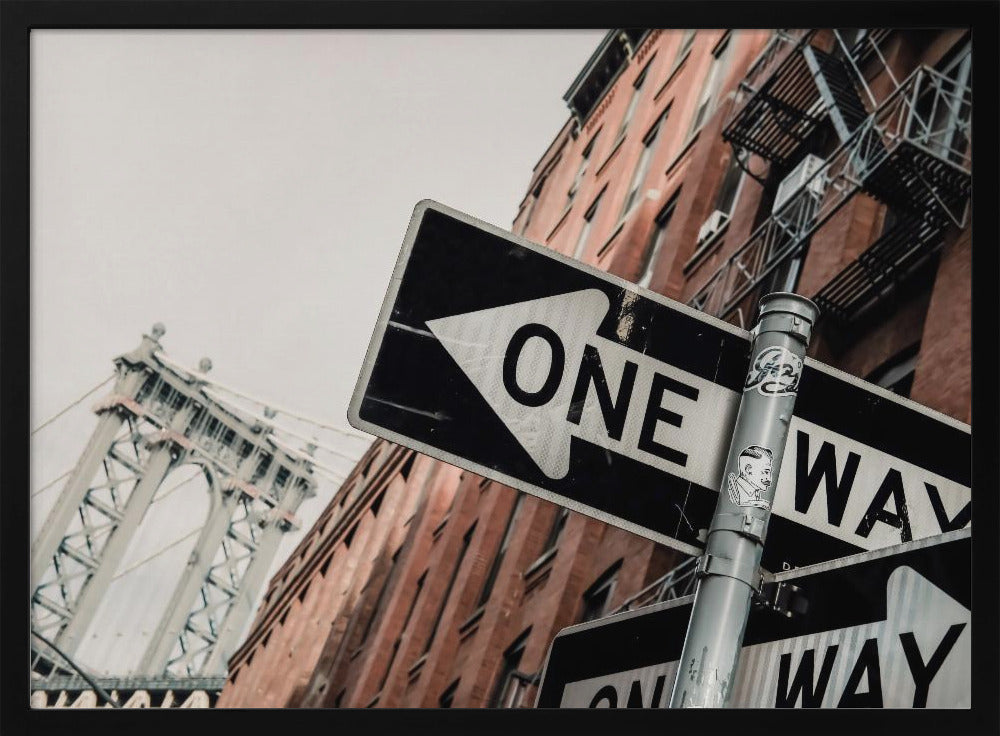 A low-angle shot of a black and white 'ONE WAY' street sign in the foreground, with a red brick building and the Manhattan Bridge visible in the background under an overcast sky, all enclosed in a silver frame. Artwork