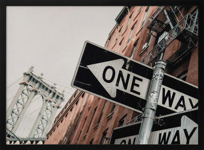 A low-angle shot of a black and white 'ONE WAY' street sign in the foreground, with a red brick building and the Manhattan Bridge visible in the background under an overcast sky, all enclosed in a silver frame. Artwork