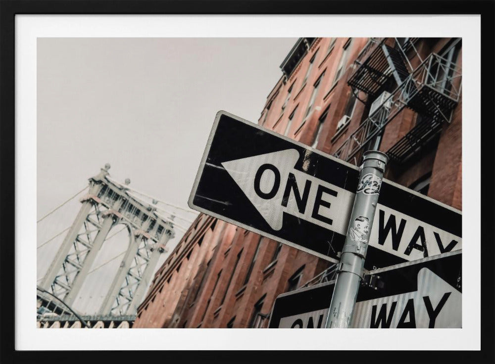 A low-angle shot of a black and white 'ONE WAY' street sign in the foreground, with a red brick building and the Manhattan Bridge visible in the background under an overcast sky, all enclosed in a silver frame. Artwork