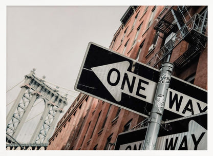 A low-angle shot of a black and white 'ONE WAY' street sign in the foreground, with a red brick building and the Manhattan Bridge visible in the background under an overcast sky, all enclosed in a silver frame. Artwork