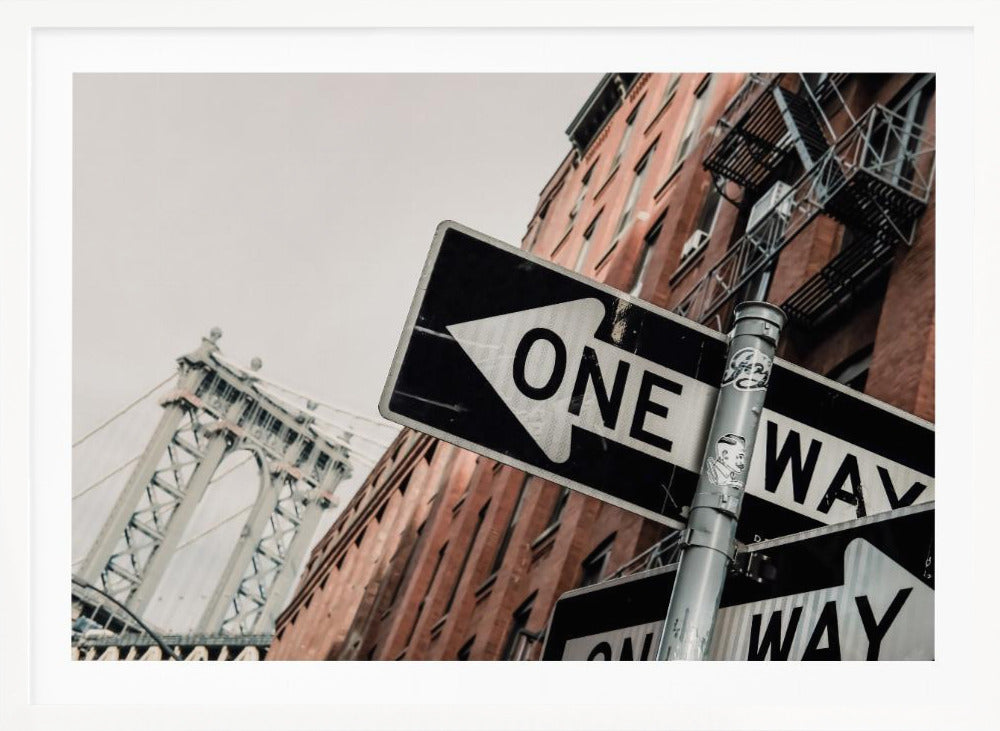 A low-angle shot of a black and white 'ONE WAY' street sign in the foreground, with a red brick building and the Manhattan Bridge visible in the background under an overcast sky, all enclosed in a silver frame. Artwork