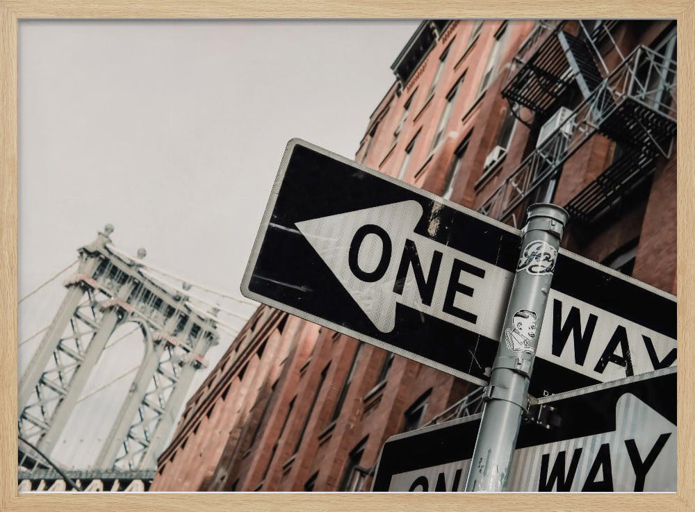 A low-angle shot of a black and white 'ONE WAY' street sign in the foreground, with a red brick building and the Manhattan Bridge visible in the background under an overcast sky, all enclosed in a silver frame. Artwork