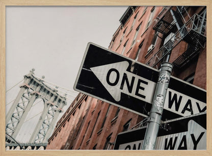 A low-angle shot of a black and white 'ONE WAY' street sign in the foreground, with a red brick building and the Manhattan Bridge visible in the background under an overcast sky, all enclosed in a silver frame. Artwork