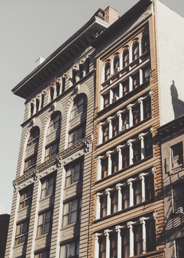 A low-angle, sunlit photograph of two ornate, multi-story historic buildings against a pale sky, framed in black. The detailed facades feature arched windows and decorative columns, with warm light creating strong shadows. Poster