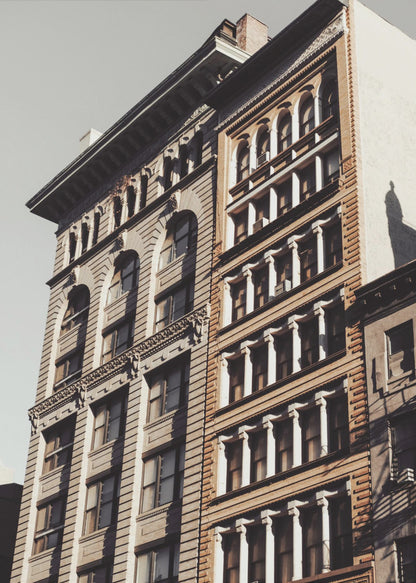 A low-angle, sunlit photograph of two ornate, multi-story historic buildings against a pale sky, framed in black. The detailed facades feature arched windows and decorative columns, with warm light creating strong shadows. Poster