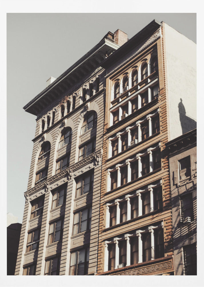 A low-angle, sunlit photograph of two ornate, multi-story historic buildings against a pale sky, framed in black. The detailed facades feature arched windows and decorative columns, with warm light creating strong shadows. Poster