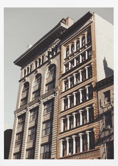 A low-angle, sunlit photograph of two ornate, multi-story historic buildings against a pale sky, framed in black. The detailed facades feature arched windows and decorative columns, with warm light creating strong shadows. Poster