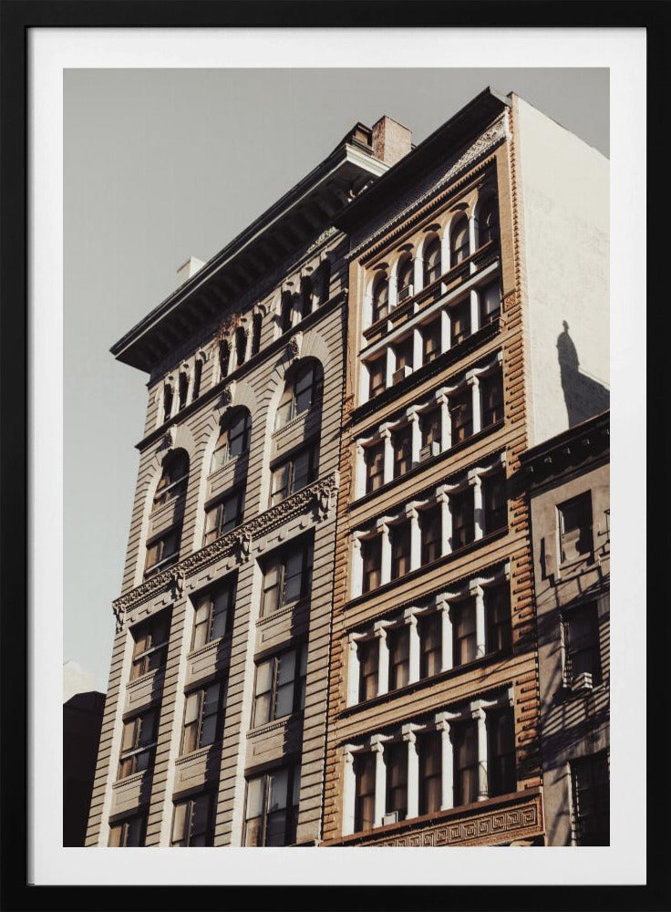 A low-angle, sunlit photograph of two ornate, multi-story historic buildings against a pale sky, framed in black. The detailed facades feature arched windows and decorative columns, with warm light creating strong shadows. Poster