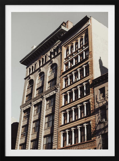 A low-angle, sunlit photograph of two ornate, multi-story historic buildings against a pale sky, framed in black. The detailed facades feature arched windows and decorative columns, with warm light creating strong shadows. Poster
