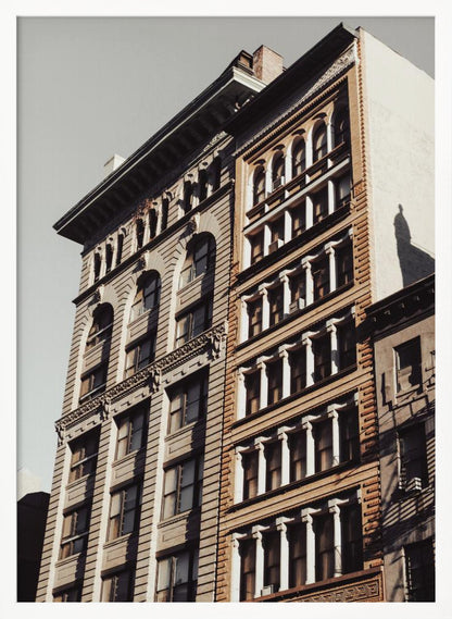 A low-angle, sunlit photograph of two ornate, multi-story historic buildings against a pale sky, framed in black. The detailed facades feature arched windows and decorative columns, with warm light creating strong shadows. Poster
