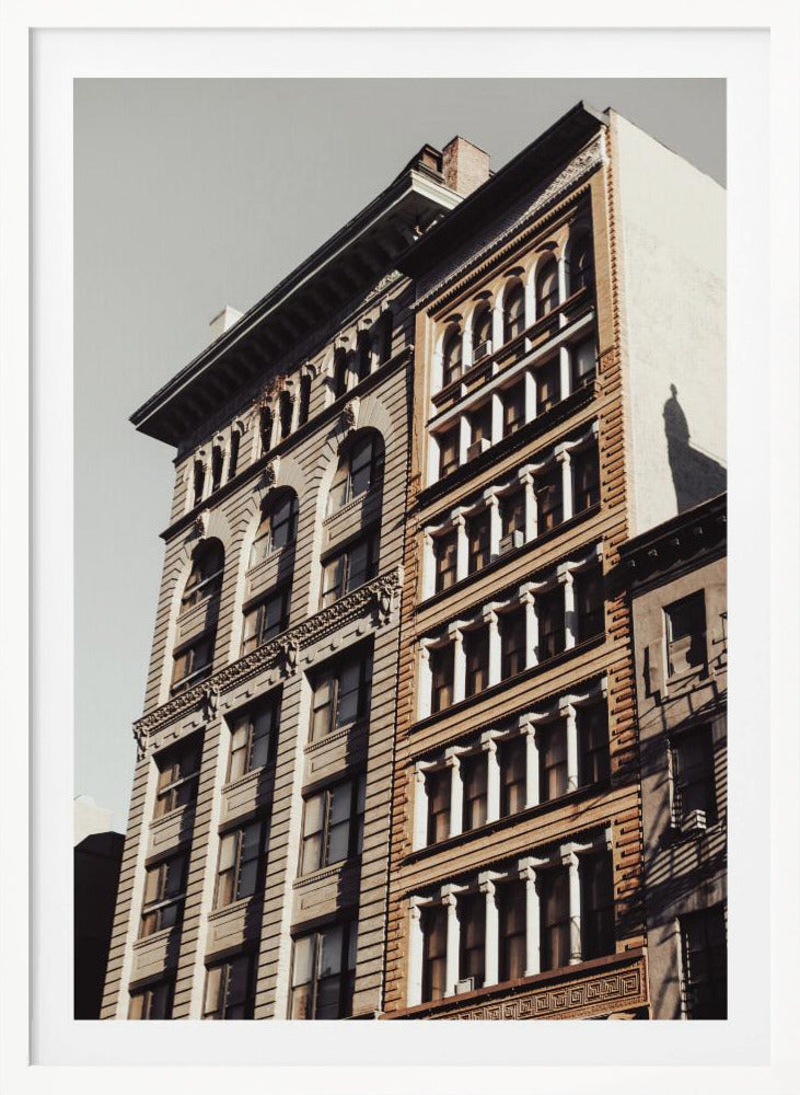 A low-angle, sunlit photograph of two ornate, multi-story historic buildings against a pale sky, framed in black. The detailed facades feature arched windows and decorative columns, with warm light creating strong shadows. Poster