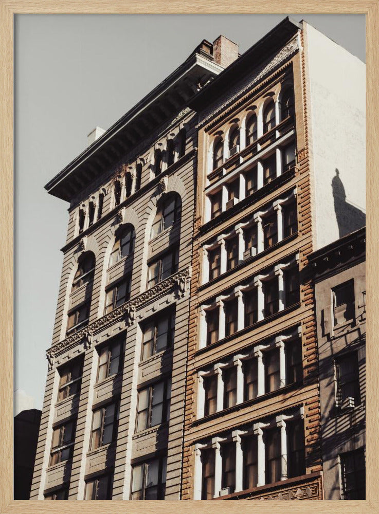 A low-angle, sunlit photograph of two ornate, multi-story historic buildings against a pale sky, framed in black. The detailed facades feature arched windows and decorative columns, with warm light creating strong shadows. Poster