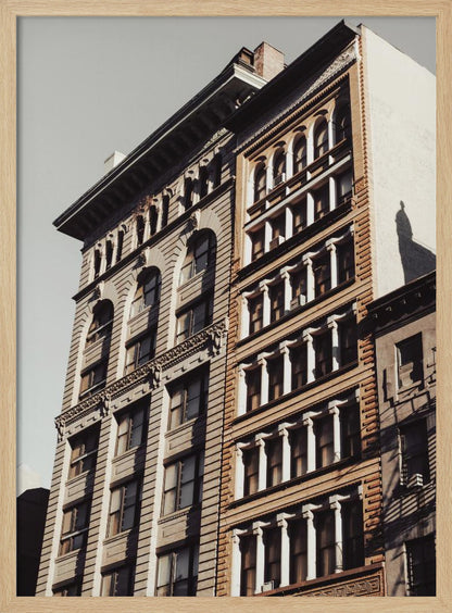A low-angle, sunlit photograph of two ornate, multi-story historic buildings against a pale sky, framed in black. The detailed facades feature arched windows and decorative columns, with warm light creating strong shadows. Poster