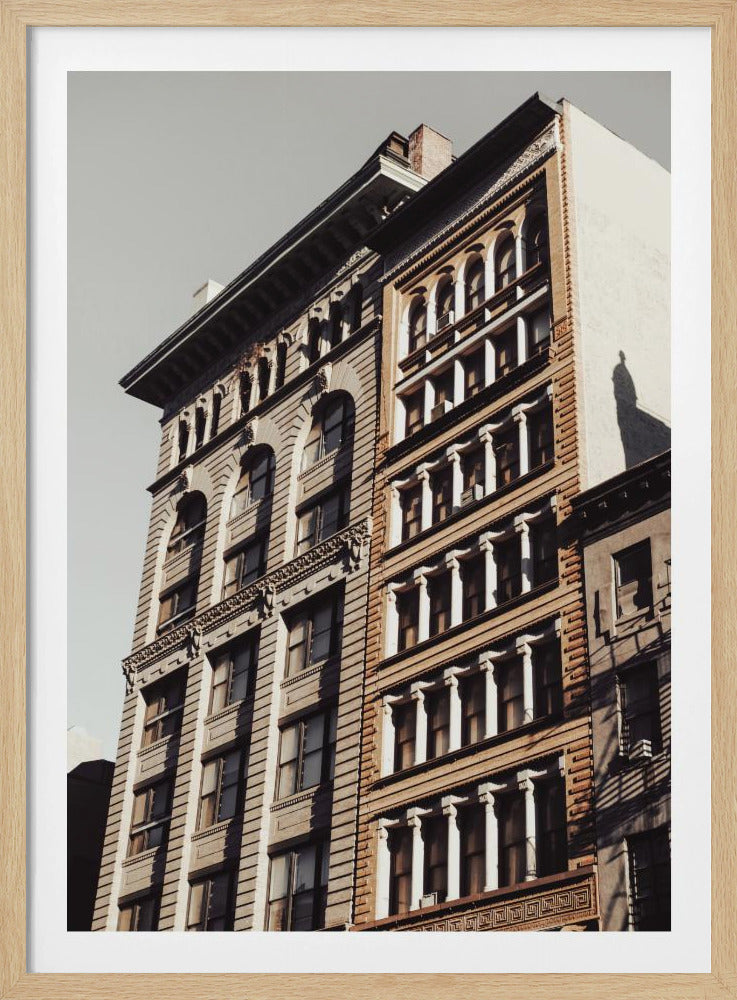 A low-angle, sunlit photograph of two ornate, multi-story historic buildings against a pale sky, framed in black. The detailed facades feature arched windows and decorative columns, with warm light creating strong shadows. Poster