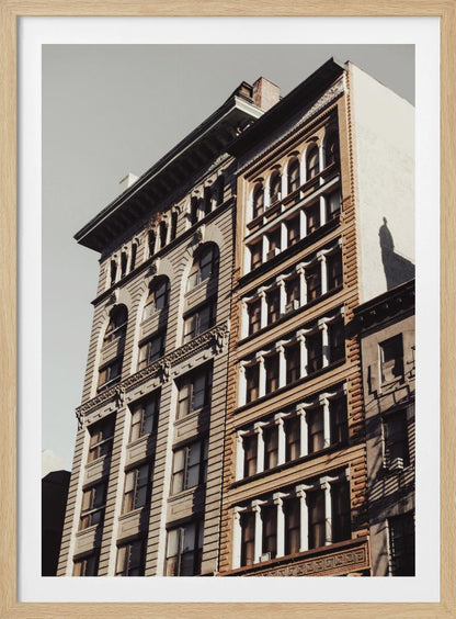 A low-angle, sunlit photograph of two ornate, multi-story historic buildings against a pale sky, framed in black. The detailed facades feature arched windows and decorative columns, with warm light creating strong shadows. Poster