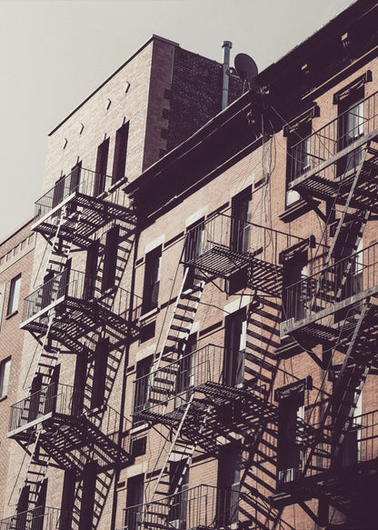 A low-angle photograph of a classic brick apartment building, with a complex pattern of black metal fire escapes casting long, dramatic shadows across the sunlit facade against a pale sky. Decor