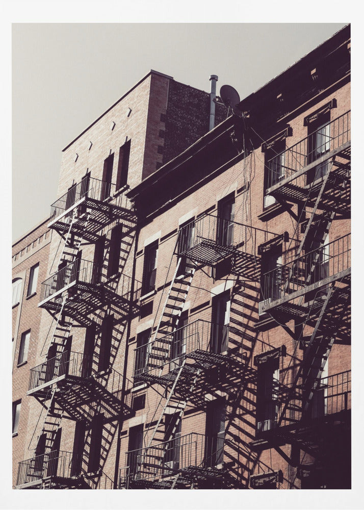 A low-angle photograph of a classic brick apartment building, with a complex pattern of black metal fire escapes casting long, dramatic shadows across the sunlit facade against a pale sky. Decor