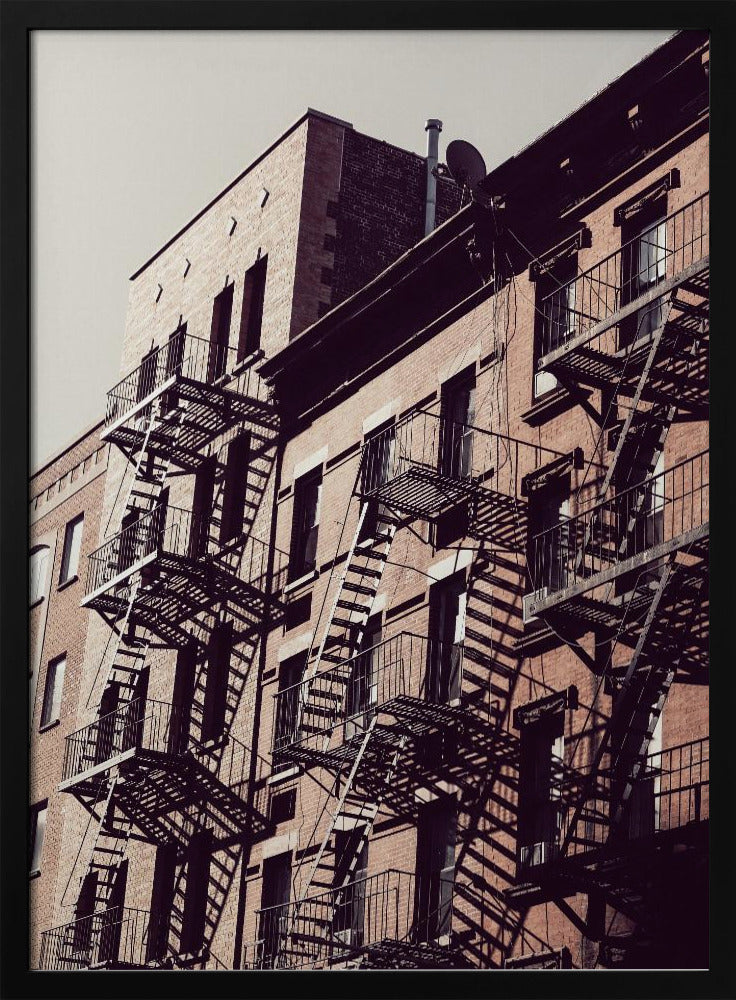 A low-angle photograph of a classic brick apartment building, with a complex pattern of black metal fire escapes casting long, dramatic shadows across the sunlit facade against a pale sky. Decor
