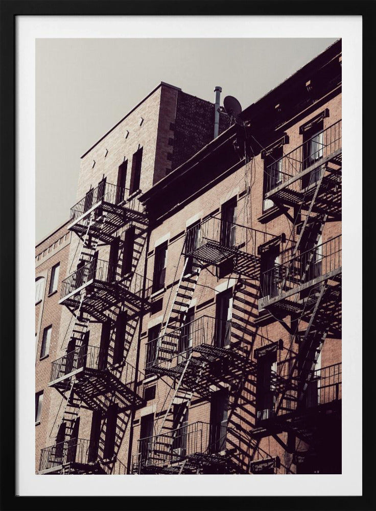 A low-angle photograph of a classic brick apartment building, with a complex pattern of black metal fire escapes casting long, dramatic shadows across the sunlit facade against a pale sky. Decor