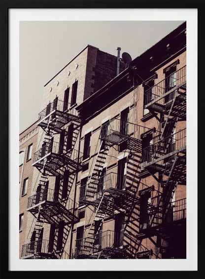 A low-angle photograph of a classic brick apartment building, with a complex pattern of black metal fire escapes casting long, dramatic shadows across the sunlit facade against a pale sky. Decor