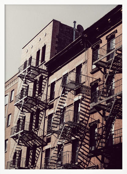 A low-angle photograph of a classic brick apartment building, with a complex pattern of black metal fire escapes casting long, dramatic shadows across the sunlit facade against a pale sky. Decor