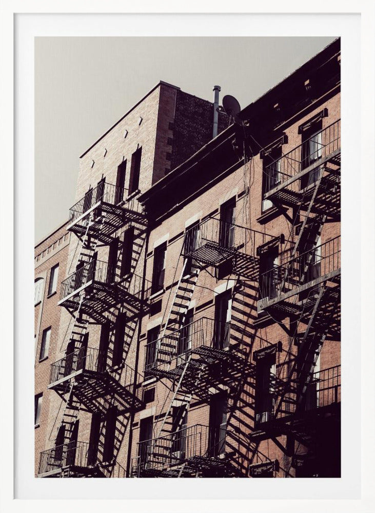 A low-angle photograph of a classic brick apartment building, with a complex pattern of black metal fire escapes casting long, dramatic shadows across the sunlit facade against a pale sky. Decor