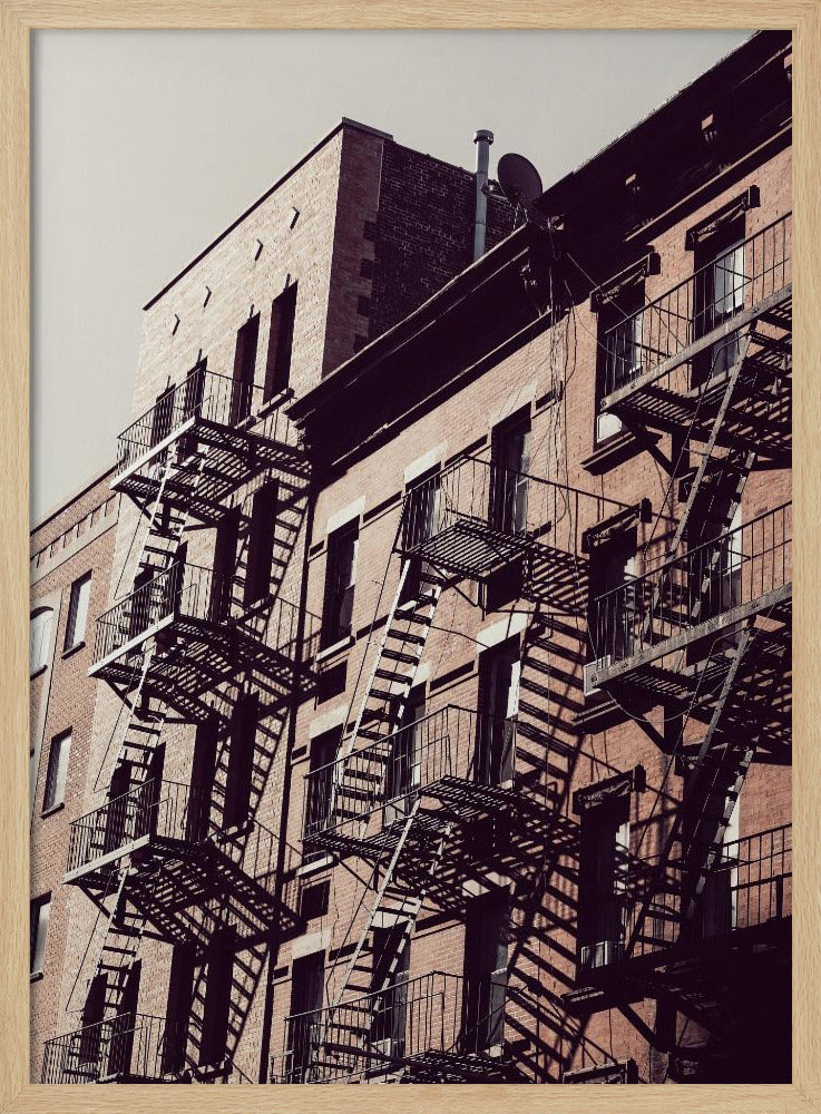 A low-angle photograph of a classic brick apartment building, with a complex pattern of black metal fire escapes casting long, dramatic shadows across the sunlit facade against a pale sky. Decor