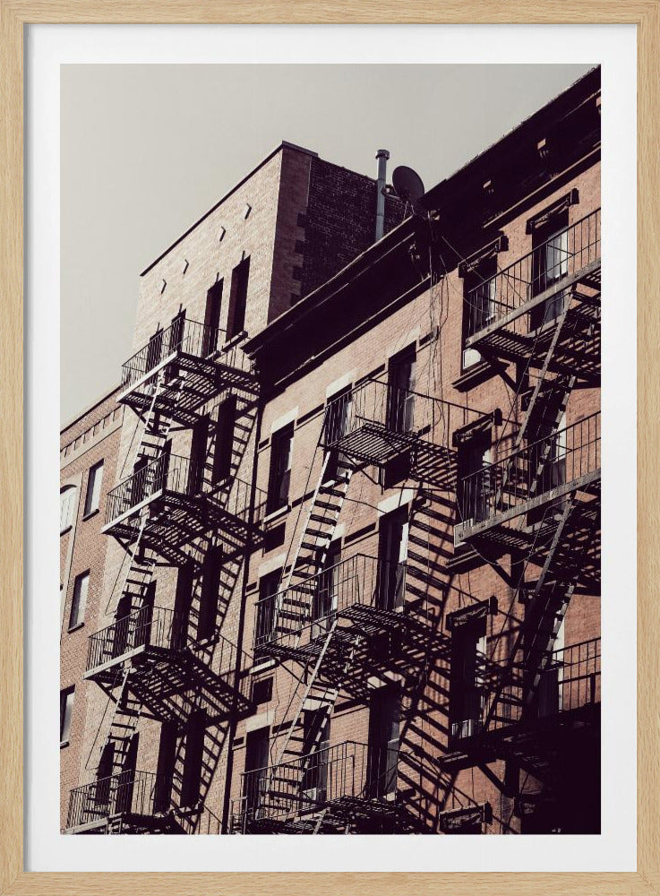 A low-angle photograph of a classic brick apartment building, with a complex pattern of black metal fire escapes casting long, dramatic shadows across the sunlit facade against a pale sky. Decor