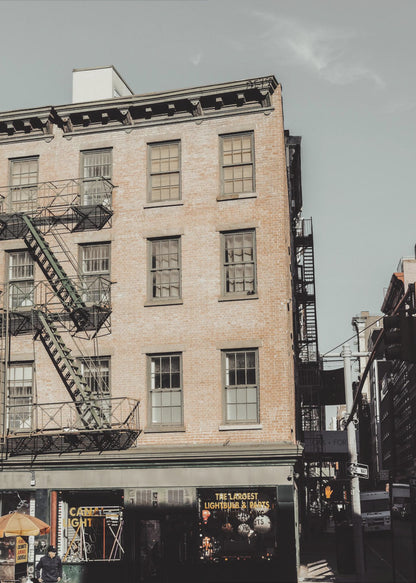 A low-angle, desaturated photograph of a classic brown brick building in a city, featuring multiple windows and a black metal fire escape zigzagging up the side. The sky is a pale, hazy blue, and the ground floor has dark storefronts. Decor