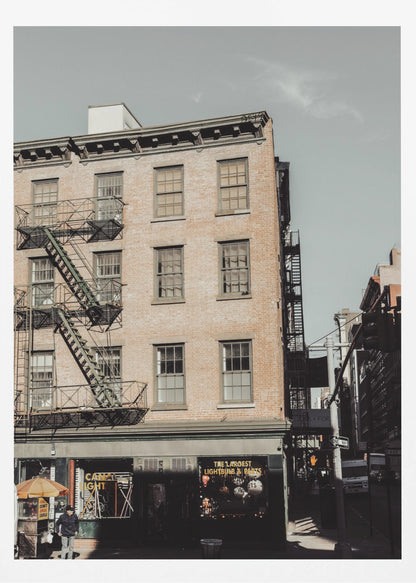 A low-angle, desaturated photograph of a classic brown brick building in a city, featuring multiple windows and a black metal fire escape zigzagging up the side. The sky is a pale, hazy blue, and the ground floor has dark storefronts. Decor