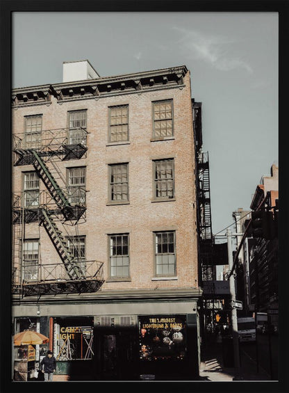 A low-angle, desaturated photograph of a classic brown brick building in a city, featuring multiple windows and a black metal fire escape zigzagging up the side. The sky is a pale, hazy blue, and the ground floor has dark storefronts. Decor