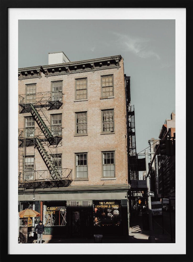 A low-angle, desaturated photograph of a classic brown brick building in a city, featuring multiple windows and a black metal fire escape zigzagging up the side. The sky is a pale, hazy blue, and the ground floor has dark storefronts. Decor