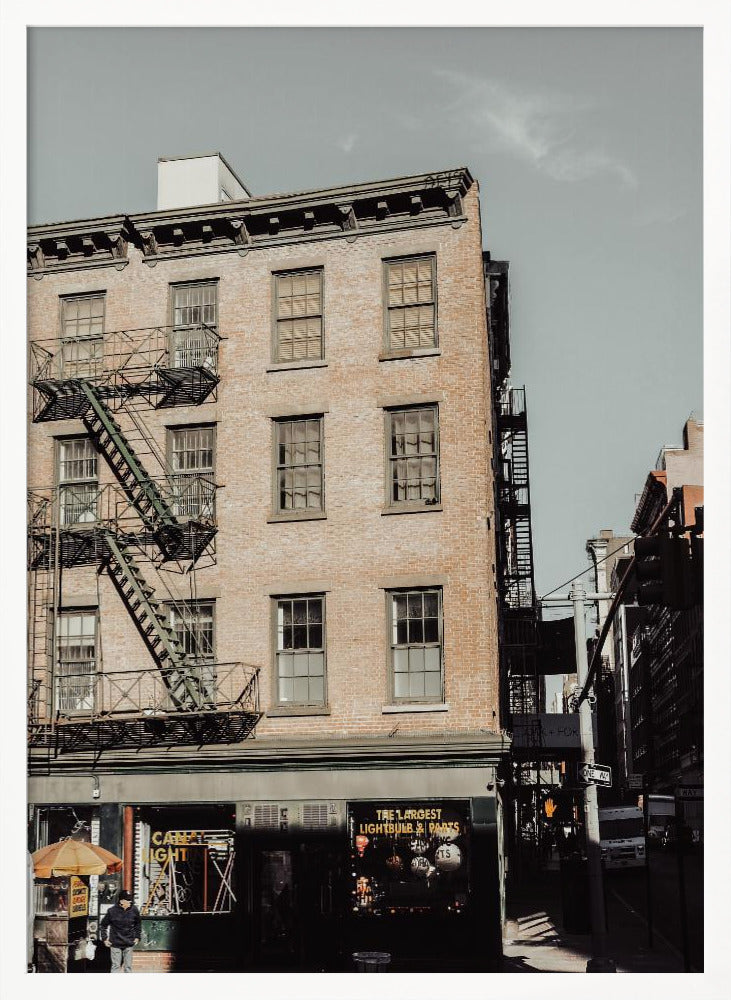 A low-angle, desaturated photograph of a classic brown brick building in a city, featuring multiple windows and a black metal fire escape zigzagging up the side. The sky is a pale, hazy blue, and the ground floor has dark storefronts. Decor