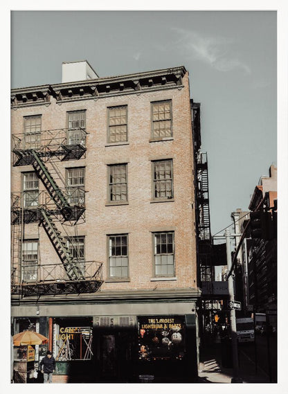 A low-angle, desaturated photograph of a classic brown brick building in a city, featuring multiple windows and a black metal fire escape zigzagging up the side. The sky is a pale, hazy blue, and the ground floor has dark storefronts. Decor