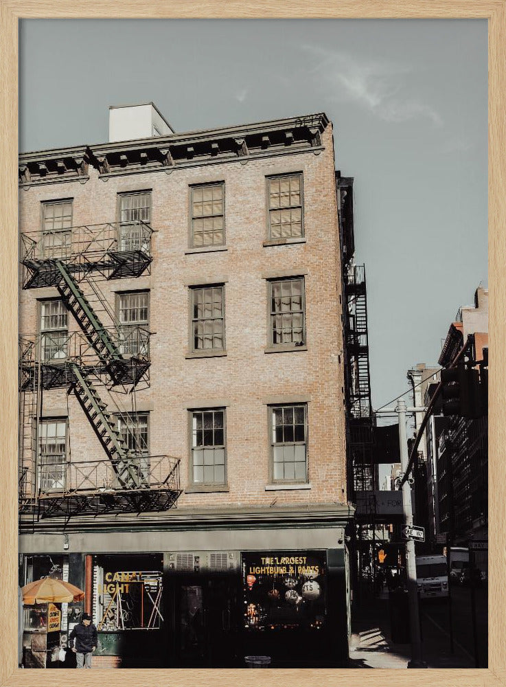 A low-angle, desaturated photograph of a classic brown brick building in a city, featuring multiple windows and a black metal fire escape zigzagging up the side. The sky is a pale, hazy blue, and the ground floor has dark storefronts. Decor
